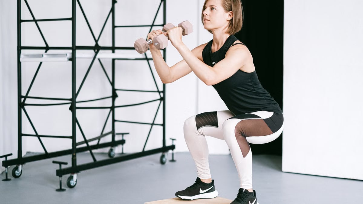 A woman in sportswear performs a squat with dumbbells indoors, embodying strength and healthy living.